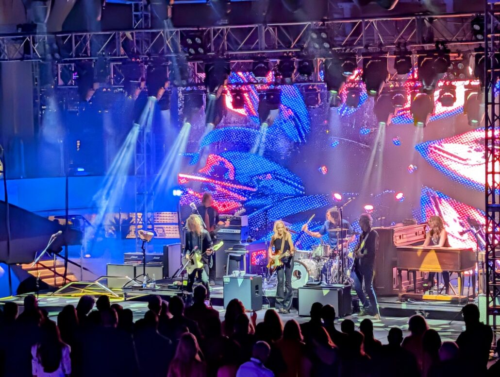 A rock band performs on a brightly lit stage with colorful LED screens in the background aboard the Star Princess. As the ship enters service, musicians play to an excited audience, while stage lights shine down, creating a lively atmosphere.