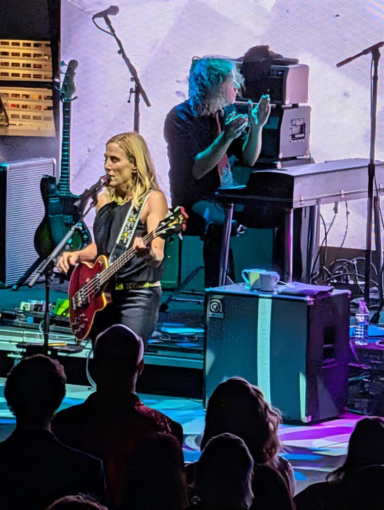 A woman plays an electric bass guitar and sings into a microphone on stage as part of the Nightly Entertainment, while a man behind her claps. Music equipment and a crowd of people are visible in the foreground and background.