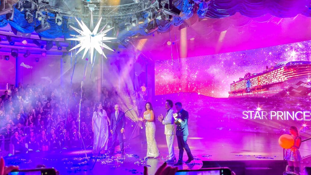 A group stands on stage beneath a large illuminated star, as the audience watches. A screen displays the Star Princess during its ship christening, with "STAR PRINCE" glowing against a purple, sparkling background.