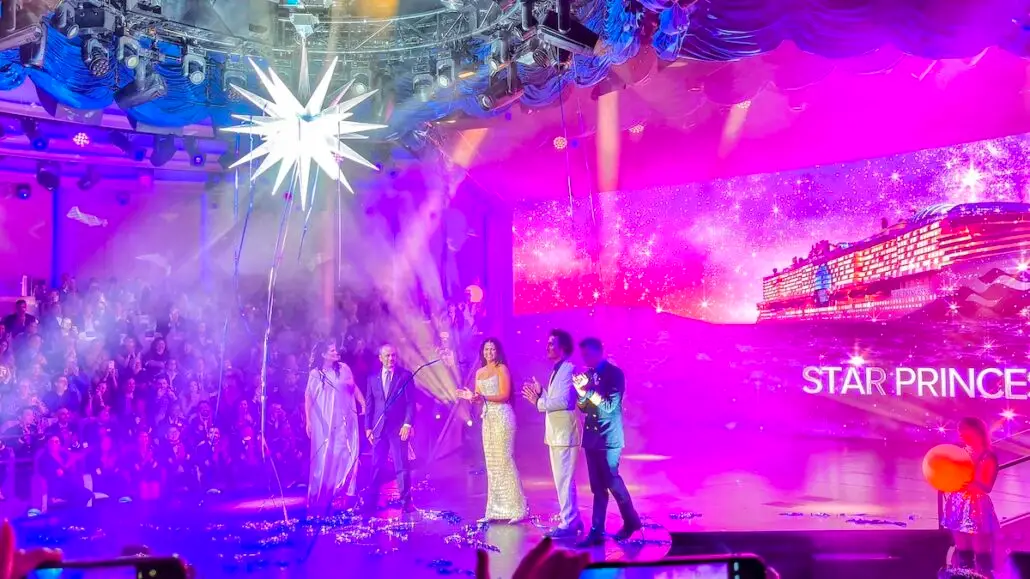 A group stands on stage beneath a large illuminated star, as the audience watches. A screen displays the Star Princess during its ship christening, with "STAR PRINCE" glowing against a purple, sparkling background.