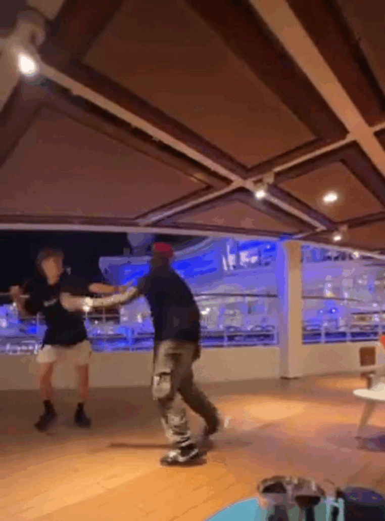 Two people are dancing together on the deck of a cruise ship at night. The area is well-lit with overhead lights, and blue lights are visible outside. There is a table with drinks in the foreground, capturing a classic cruise vibe for live streamers.