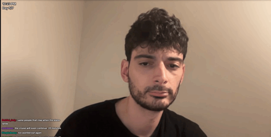 A man with short, curly dark hair and a trimmed beard looks at the camera with a neutral expression. Wearing a black shirt, he sits against a plain beige wall as live streamers’ chat messages appear in the bottom left corner.
