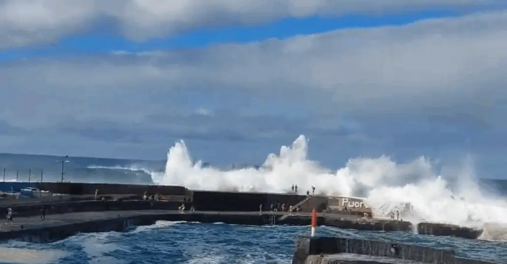 Large wave surges crash against a concrete pier under a partly cloudy sky, sending white spray high into the air. People, possibly cruise guests, stand along the pier, and "Puer" is visible on a wall along the rough sea of the Tenerife coast.