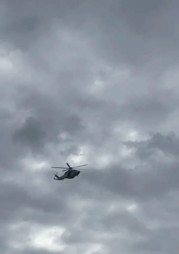 A helicopter is flying under a cloudy sky, appearing dark against the overcast Atlantic background as it conducts a search for a man overboard. No other objects are visible.
