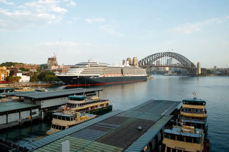 A large cruise ship is docked in a harbor near a city with several ferries in the foreground and a large steel arch bridge in the background under a partly cloudy sky.