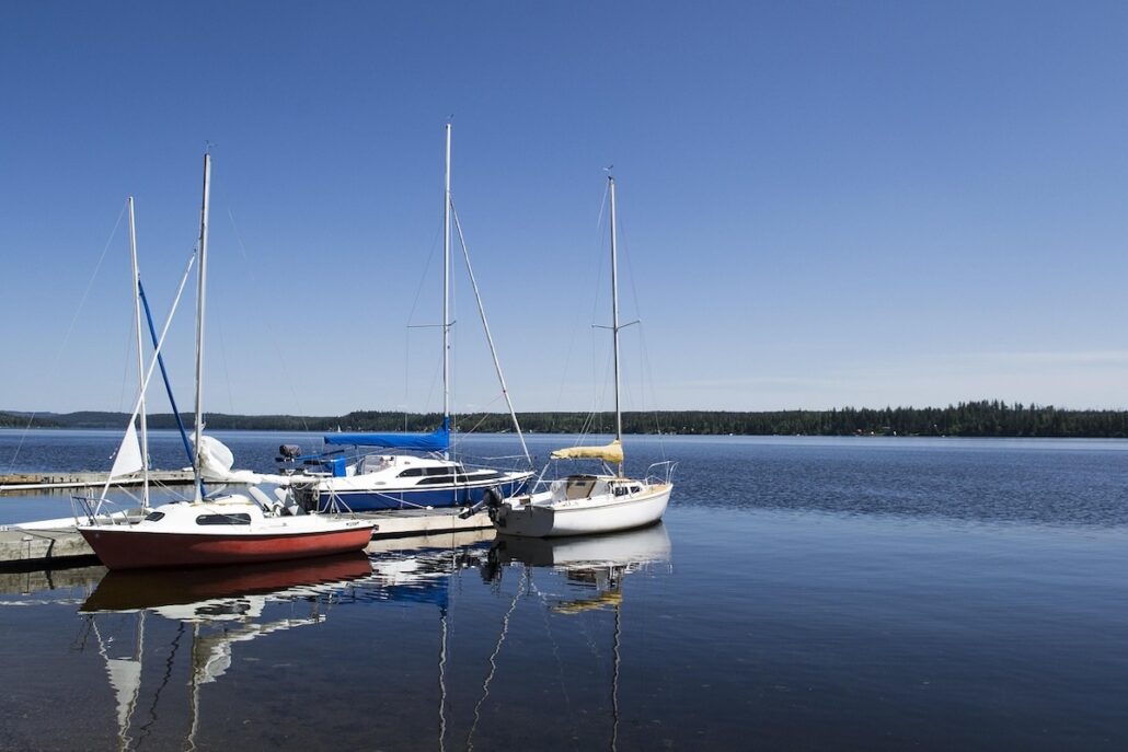 Three small sailboats are moored at a wooden dock on a calm lake under a clear blue sky, embodying responsible cruising with the serene forested shoreline visible in the distance.