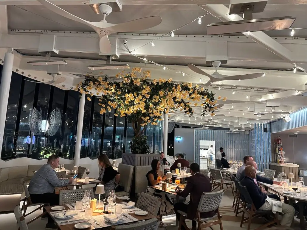 People dine at tables in a modern restaurant aboard Celebrity Xcel, with large windows, ambient lighting, and a ceiling decorated with yellow flowers and ceiling fans. The relaxed atmosphere invites conversation worthy of a glowing cruise review.