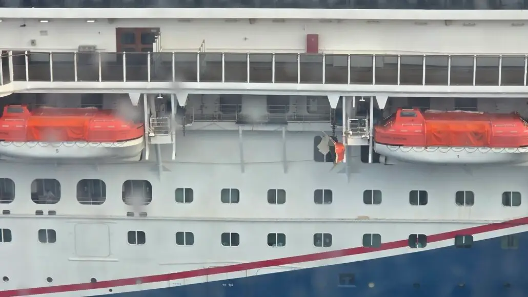 A close-up view of the side of a Carnival ship shows two orange-covered lifeboats and a crew member in an orange uniform standing near an open hatch. The ship’s exterior is white with blue and red accents.