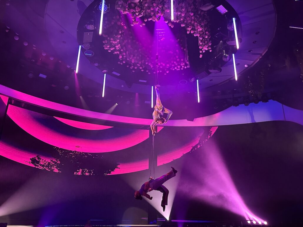 Two acrobats perform on a vertical pole on stage under purple and pink lighting aboard Celebrity Xcel. One is suspended upside down while the other holds above, with light beams and a curved stage design enhancing this memorable Celebrity Cruises show.