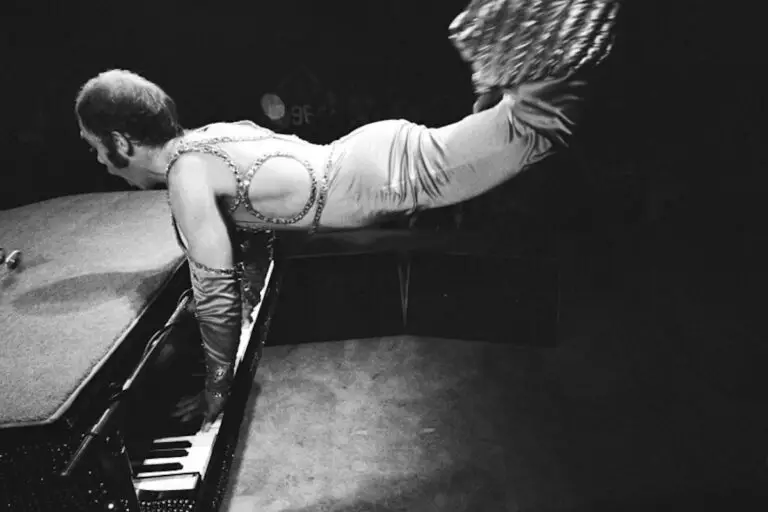 A person in a sequined bodysuit and gloves balances horizontally above a piano, supporting themselves with their arms on the keys, during the Norwegian Luna Elton John Tribute Show stage performance.
