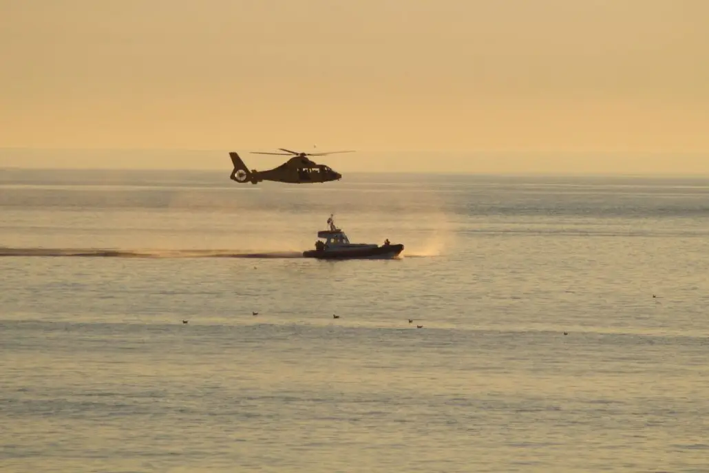 A helicopter hovers low above the Atlantic near a small boat, creating ripples and mist on the water under a golden sky during a search for a man overboard. Several birds float in the water nearby.