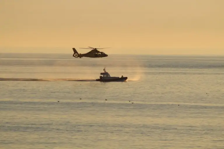 A helicopter hovers low above the Atlantic near a small boat, creating ripples and mist on the water under a golden sky during a search for a man overboard. Several birds float in the water nearby.