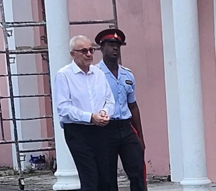 An older man in a white shirt walks with his hands together, escorted by a uniformed officer in front of pink walls and white columns; a metal scaffold is visible on the left amid reports of a sexual assault allegation at Castaway Cay.