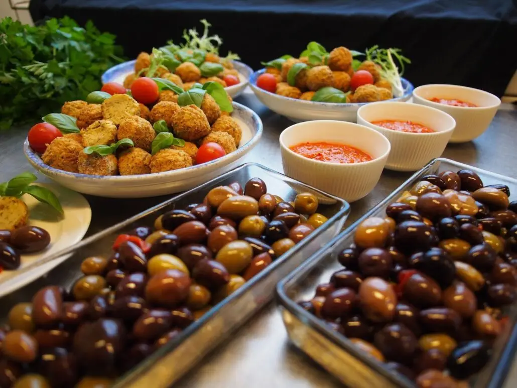 Bowls of falafel garnished with cherry tomatoes and basil, three bowls of red dipping sauce, and trays of assorted olives are arranged on a metal surface—perfect for first-time visitors Spain seeking authentic flavors.