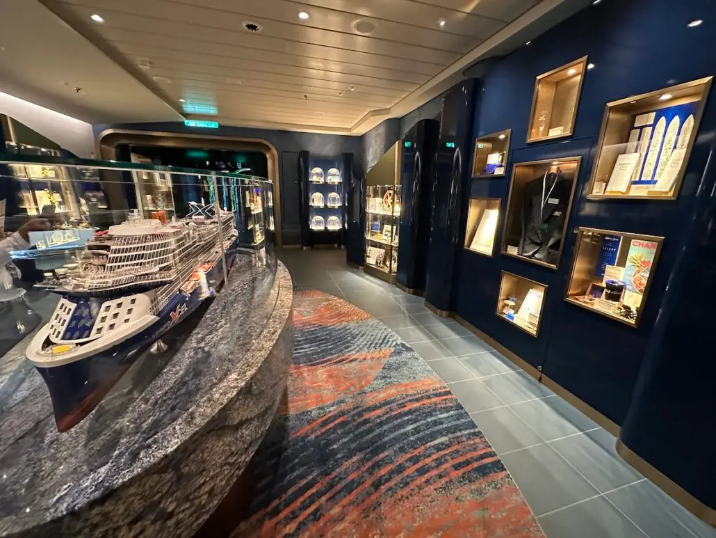 A well-lit display room with glass cases showing ship-related memorabilia, including a detailed Celebrity Xcel model ship on a marble counter and various items on shelves along the dark blue walls.