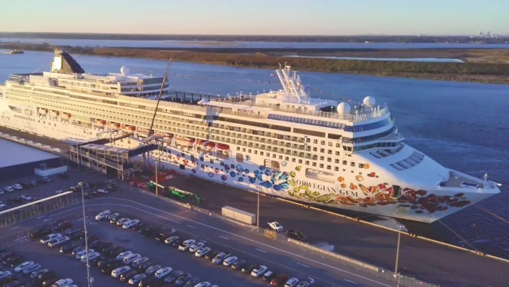 A large white Norwegian Cruise Line ship with colorful decorations is docked at a port terminal. Rows of parked cars fill the foreground, and a river with grassy banks is visible in the background.