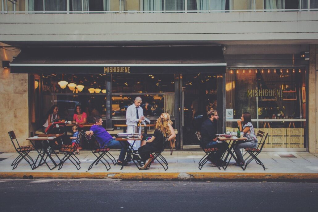Several people sit at outdoor tables in front of a restaurant while a waiter serves one table. The restaurant has large windows and a black awning with the sign "Mishiguene"—a vibrant spot for First-Time Visitors Spain should add to their list.
