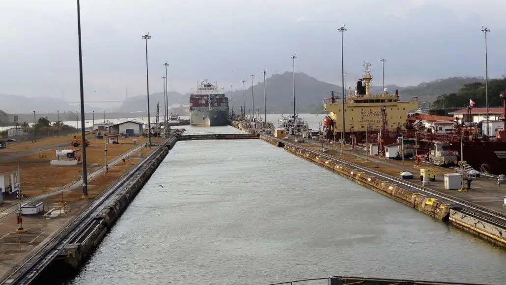 A large cargo ship moves through a canal lock system, with another vessel docked on the right—an impressive scene often encountered during Caribbean Sailings. The area is surrounded by water, buildings, and hills beneath a cloudy sky.