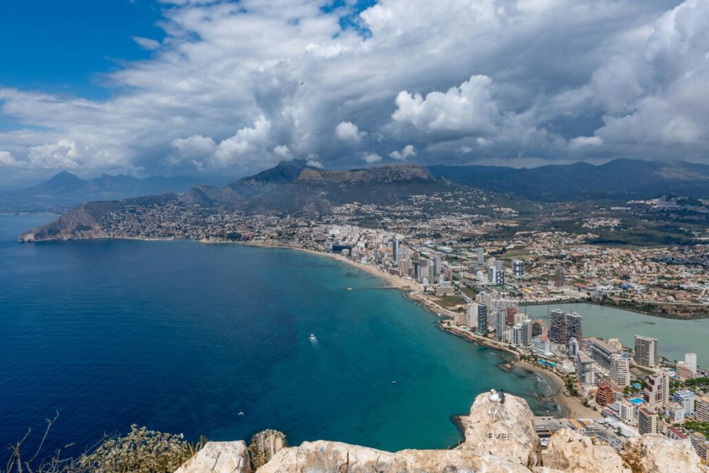 A coastal city with high-rise buildings lines a sandy shoreline next to bright blue water. Perfect for first-time visitors, Spain travel offers stunning views with hills and mountains in the background and rocks visible in the foreground.