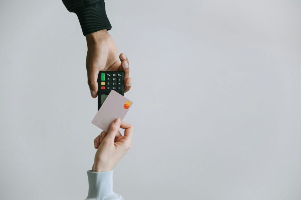 A person holds a payment terminal while another holds a credit card, preparing to make a contactless payment—a common scene for first-time visitors Spain may encounter during their travels. The background is plain and light gray.