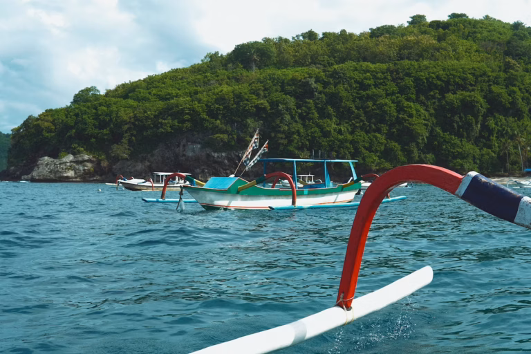 Wooden boats float on blue water near a forested island shoreline. Part of a boat’s red and white outrigger is visible in the foreground. The sky is mostly cloudy.