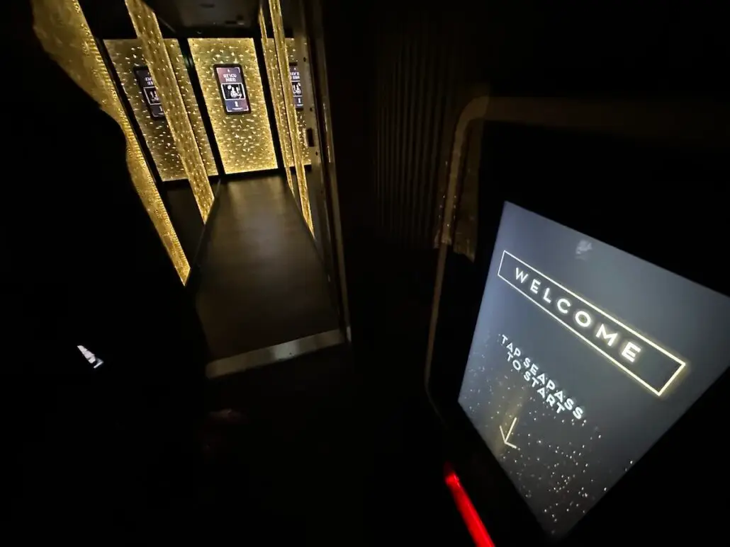 A dimly lit hallway with yellow lights on the walls and a touchscreen monitor in the foreground displaying “WELCOME” hints at the high-tech ambiance of Celebrity Xcel, elevating your Celebrity Cruises experience from the moment you arrive.