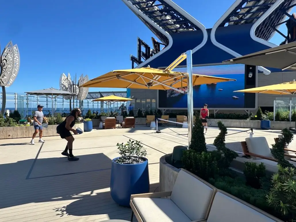 People play pickleball on a rooftop court under large yellow umbrellas aboard Celebrity Xcel, with modern architecture and ocean in the background. Potted plants and lounge seating add to the upscale Celebrity Cruises experience.