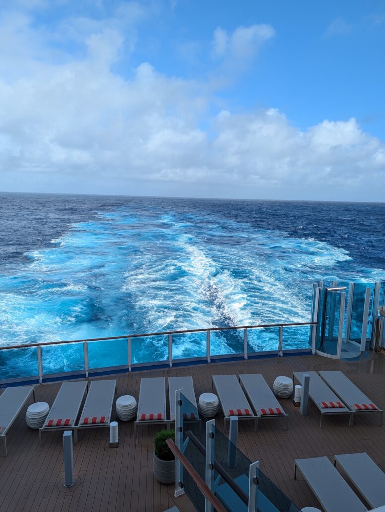 View from the Star Princess cruise ship showing empty lounge chairs on the deck and a wake trailing behind in the blue ocean under a partly cloudy sky, perfect for those seeking quiet spaces to relax.