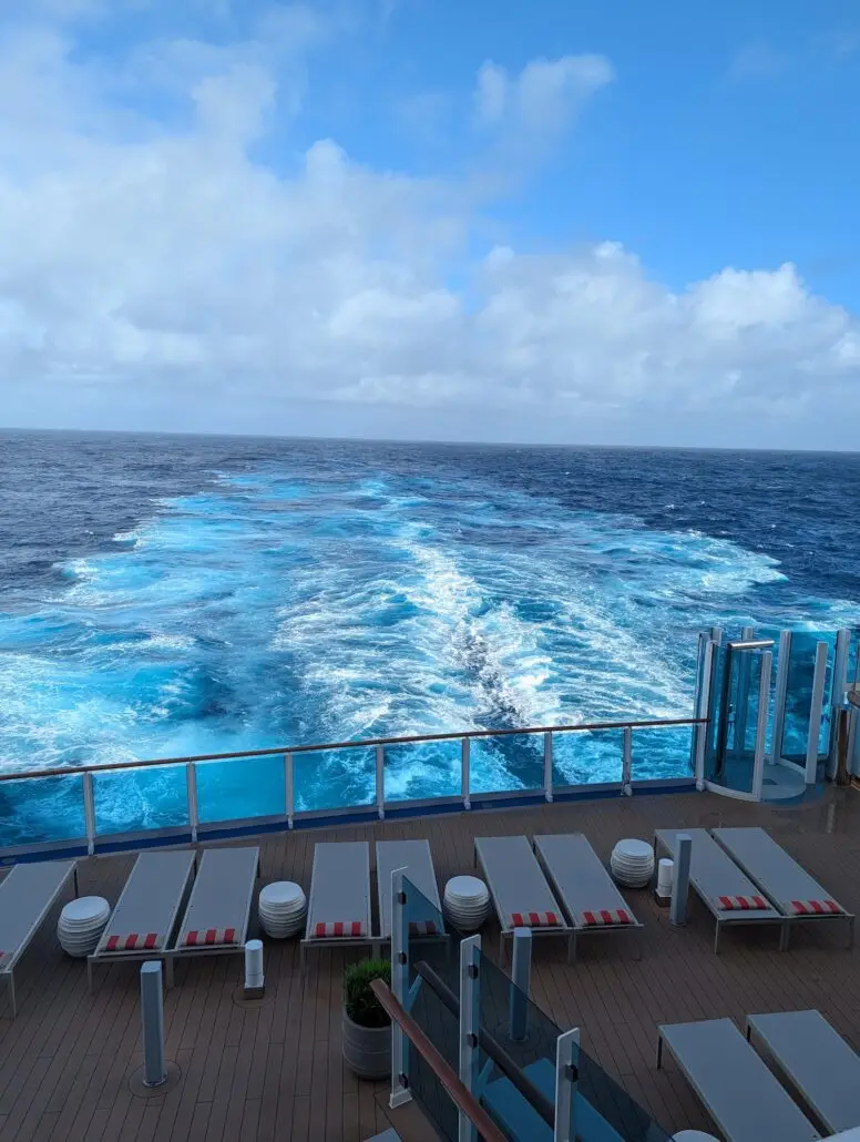 View from the Star Princess cruise ship showing empty lounge chairs on the deck and a wake trailing behind in the blue ocean under a partly cloudy sky, perfect for those seeking quiet spaces to relax.