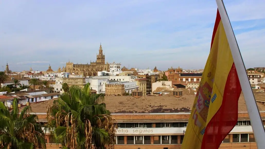 A cityscape view of Seville, Spain, with the Giralda tower and Seville Cathedral in the background, palm trees in the foreground, and a Spanish flag on the right—perfect inspiration for first-time visitors Spain or anyone seeking Spain travel tips.