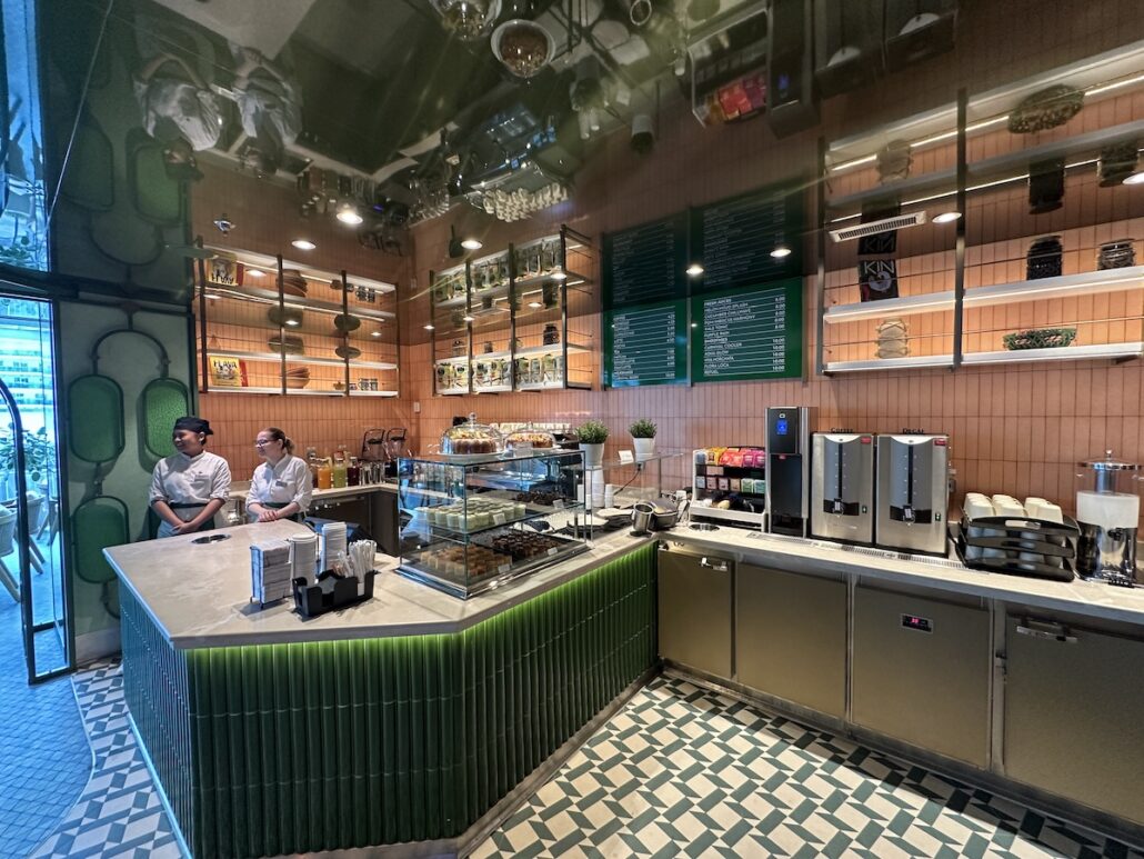 A modern café interior with two people talking behind a counter evokes the relaxed elegance found on Celebrity Xcel. Shelves with jars and decor line the walls, pastries fill a glass display case, and coffee machines sit to the right on green and white tiled floors.