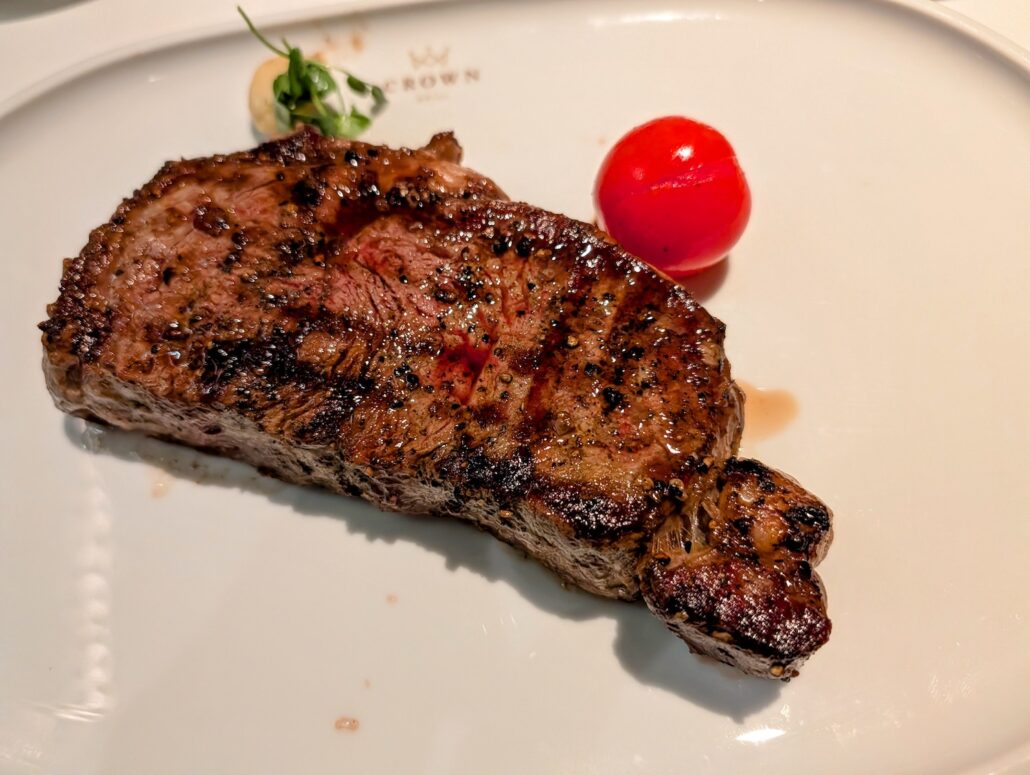 A grilled steak with pepper seasoning sits on a white plate next to a small round red tomato and a garnish of greens, perfect for enjoying after an evening of nightly entertainment at Princess Cay.