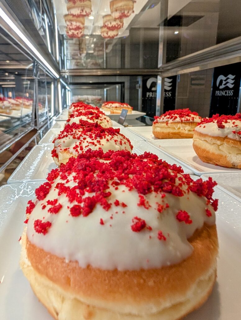 Rows of round donuts with white icing and red crumbles are displayed on a tray in a glass case at Princess Cay; their reflection is visible in the mirrored surface above and behind them, tempting anyone who loves delicious food.