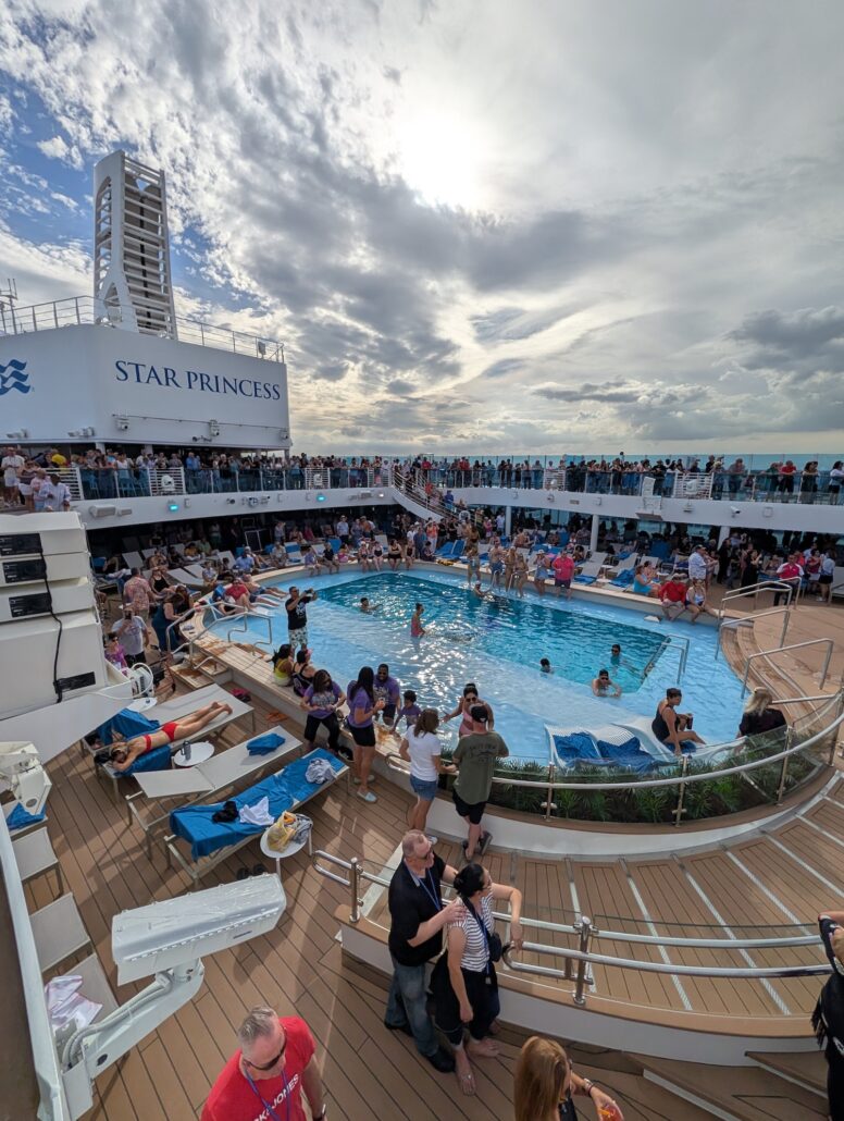 A crowd gathers around the swimming pool on the deck of the Star Princess cruise ship under a cloudy sky, with some sunbathers relaxing and others enjoying activity in the pool before heading to nightly entertainment.