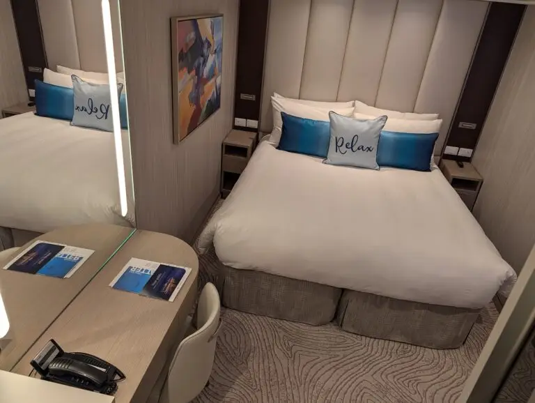 A neatly made bed with white linens and two blue "Relax" pillows welcomes you to a compact, modern Star Princess room. Beside the bed are a desk, chair, phone, brochures, and large mirror—perfect for enjoying quiet spaces after exploring dining options.
