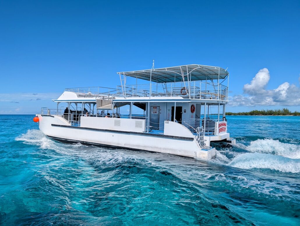 A white double-deck boat cruises through the clear turquoise water near Princess Cay under a bright blue sky. A few people are on board, enjoying the scenic shoreline lined with trees in the background.