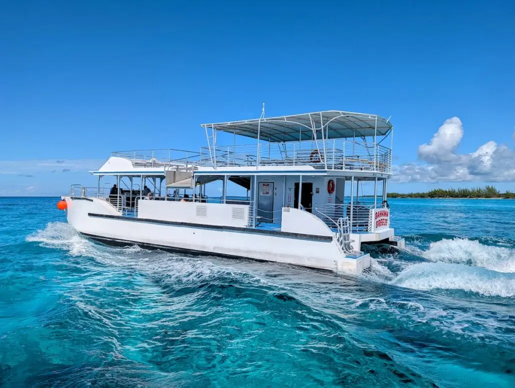 A white double-deck boat cruises through the clear turquoise water near Princess Cay under a bright blue sky. A few people are on board, enjoying the scenic shoreline lined with trees in the background.