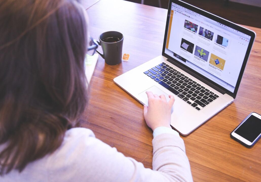 A person using a laptop at a wooden table, browsing a website with Spain travel tips and articles. A smartphone, a cup of tea, and papers are on the table. The person’s hand is on the laptop trackpad.