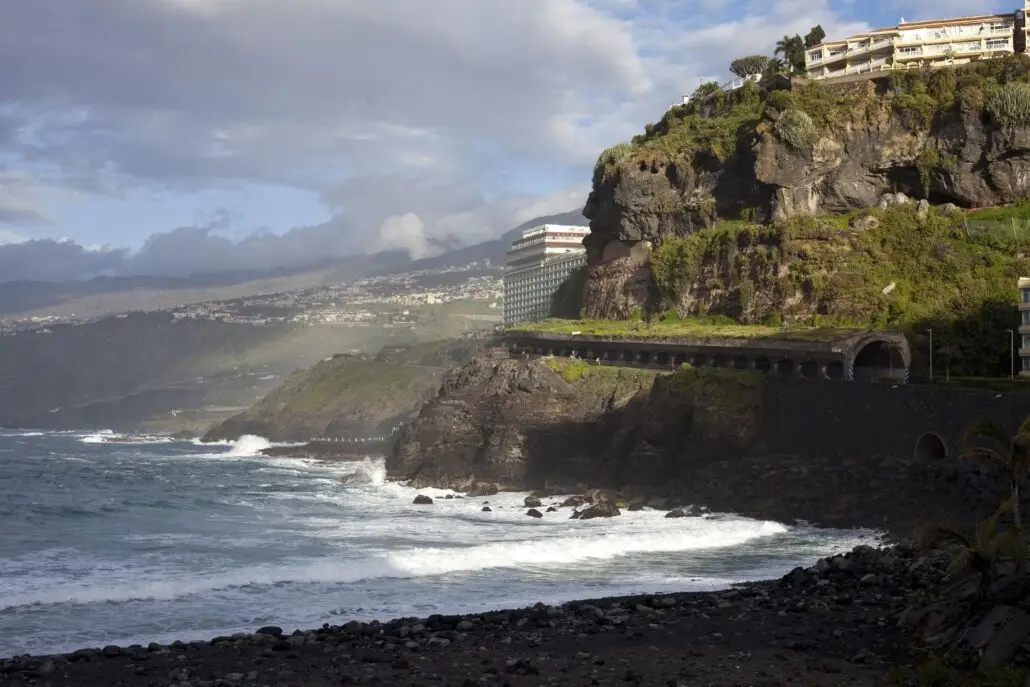 A rocky Tenerife coast with wave surges crashing against dark cliffs, a tunnel built into the cliffside, and buildings perched above. Cloudy skies and distant mountains complete the dramatic scene.