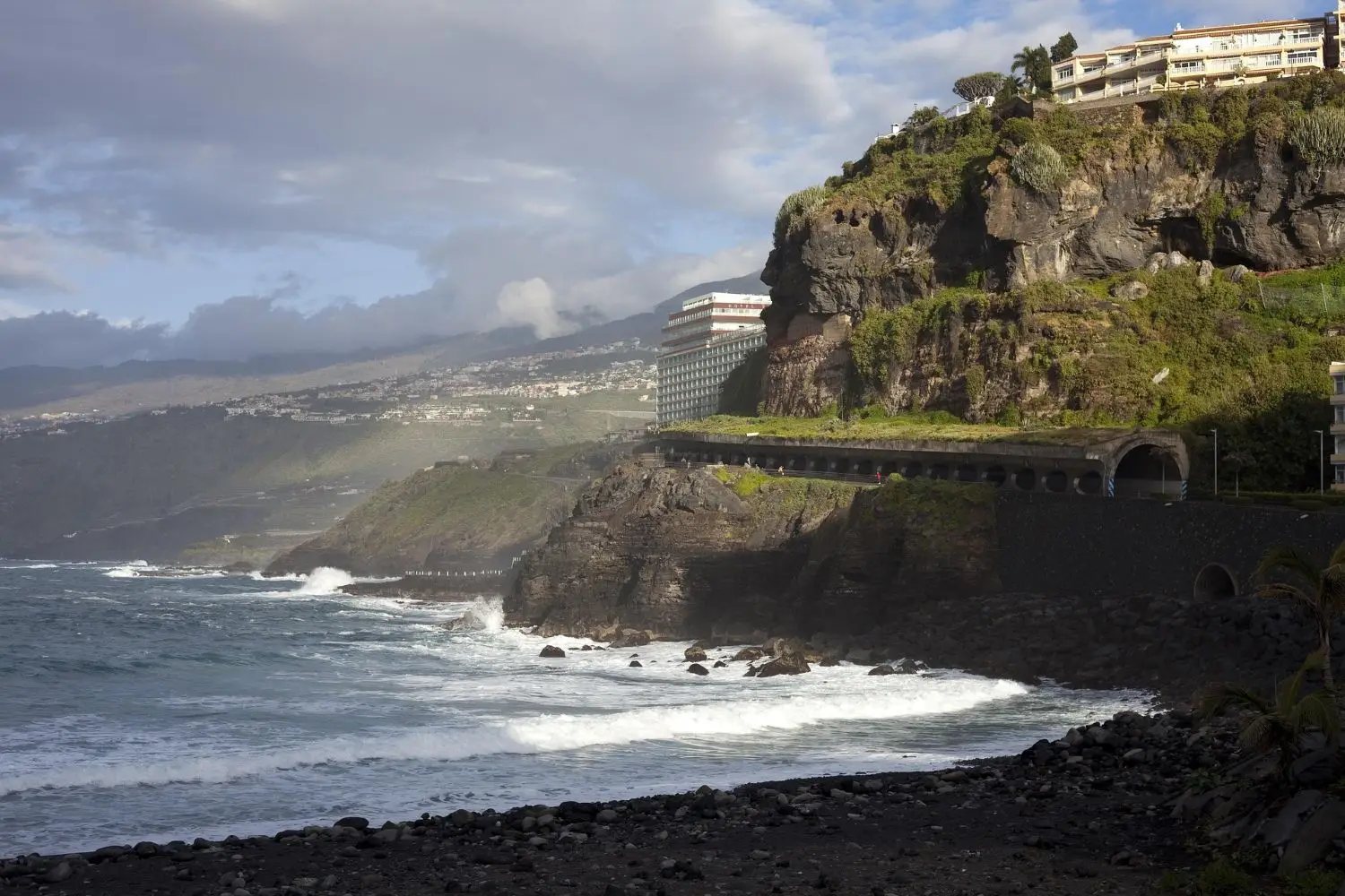 A rocky Tenerife coast with wave surges crashing against dark cliffs, a tunnel built into the cliffside, and buildings perched above. Cloudy skies and distant mountains complete the dramatic scene.