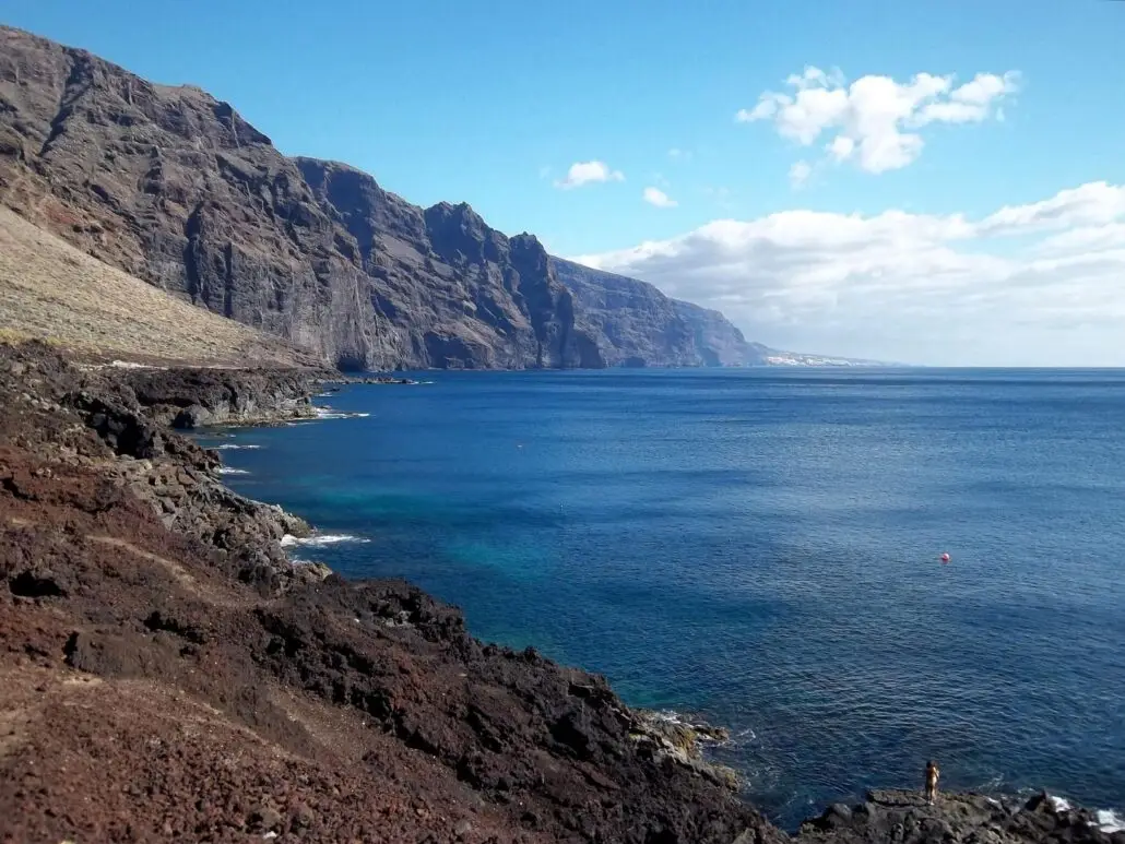 Rocky Atlantic coastline with steep cliffs on the left, meeting a deep blue ocean under a mostly clear sky with some clouds. A small buoy floats in the water near the shore, possibly marking a search area for a man overboard.