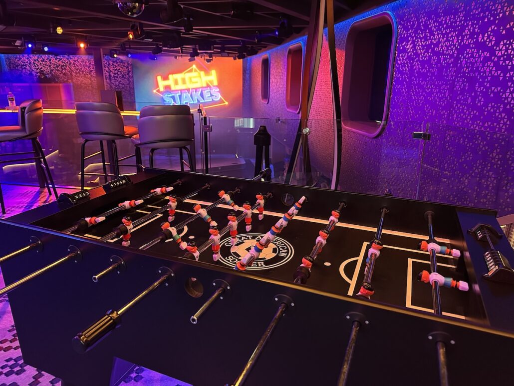 A foosball table is in the foreground of a dimly lit room with purple and orange lighting aboard Celebrity Xcel. In the background, bar stools, a table, and a neon sign reading “HIGH STAKES” evoke the vibrant nightlife of Celebrity Cruises.