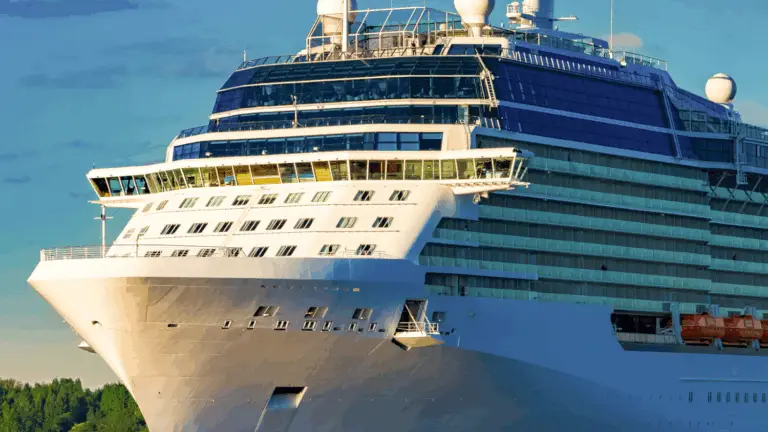 The front view of a large cruise ship with multiple decks and windows, docked near green trees under a partly cloudy sky, hints at bustling onboard logistics and the excitement of a cruise ship casino awaiting guests.