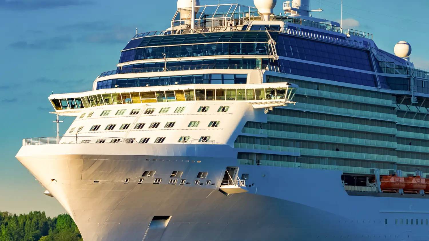 The front view of a large cruise ship with multiple decks and windows, docked near green trees under a partly cloudy sky, hints at bustling onboard logistics and the excitement of a cruise ship casino awaiting guests.