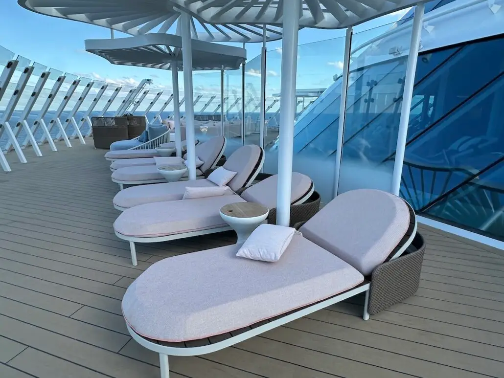 Several lounge chairs with cushions and small side tables line the deck of Celebrity Xcel under white canopies, with glass railings and a view of the sky in the background—a perfect spot for your Celebrity Cruises experience.