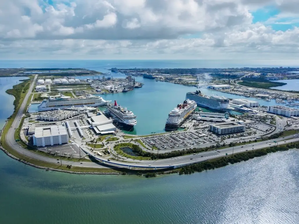 Aerial view of Port Canaveral, one of the busiest cruise ports, with several cruise ships docked, surrounded by parking lots, buildings, and water channels on a sunny day with scattered clouds.