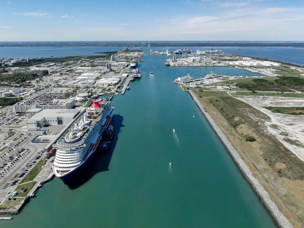 A large cruise ship is docked at Port Canaveral, one of the world's busiest cruise ports, near an industrial area and waterways, with several small boats in the channel and buildings and roads stretching into the distance.