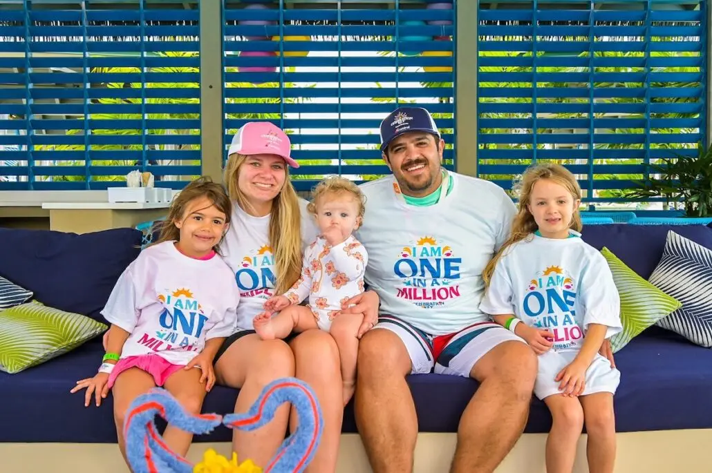 A family of five sits on a blue couch. The adults and two children wear matching white shirts with colorful "I Am One in a Million" text. A baby sits in the center wearing a patterned onesie. Blue shutters are in the background.