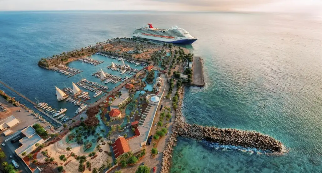 A large Carnival cruise ship is docked at the Ensenada Cruise Port, with numerous sailboats and palm trees lining the marina. The marina extends into the blue ocean, surrounded by breakwaters and landscaped pathways.