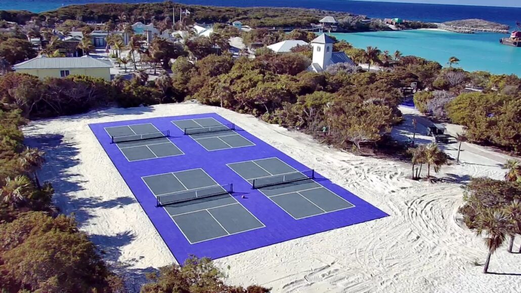 Aerial view of four tennis courts with blue surfaces and black nets, surrounded by sand and trees, near buildings and turquoise water on an island.